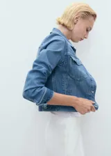 Chaqueta de jean azul de corte crop, con cuello camisero, cierre frontal de botones metálicos, bolsillos con solapa en el pecho y mangas largas con puños abotonados.