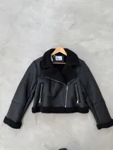 Campera tipo gamulan de cuero sintético negro, con interior y detalles en corderito sintético al tono. Presenta cierre frontal cruzado, bolsillos laterales con cierre y solapas amplias.