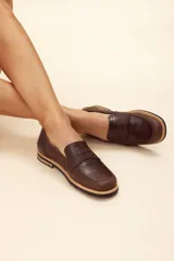 Mocasín de cuero vacuno en color marrón, con diseño bitono que combina texturas lisas y con grabado tipo cocodrilo en el empeine. Cuenta con detalle de estribo clásico y suela plana con vira en contraste.