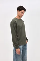 Remera de manga larga color verde oliva, confeccionada en algodón pesado con textura suave. Presenta un bolsillo frontal en el pecho con un pequeño bordado de la marca y puños acanalados en las mangas.