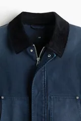 Campera worker corta de sarga de algodón en color azul oscuro, con cuello de pana negro en contraste. Cuenta con cierre frontal oculto por solapa con botones a presión y grandes bolsillos frontales tipo parche.