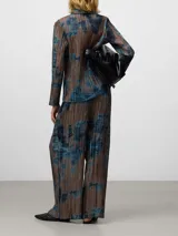 Pantalón ancho de tiro alto con efecto plisado (crinkle) y estampado abstracto en tonos marrón topo y azul. Presenta un diseño cruzado en el frente con cierre de botón y cintura elástica.