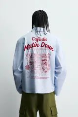 Camisa de algodón de corte regular, color celeste con finas rayas blancas verticales. Presenta un bordado en color rojo en el pecho con la frase "Café du Matin Doux".