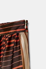 Falda midi de tiro alto con bajo asimétrico. Presenta un diseño patchwork con bloques de diferentes estampados, incluyendo rayas verticales en verde lima, rayas diagonales en marrón oscuro y naranja, y un estampado a cuadros en tonos verdes y marrones.