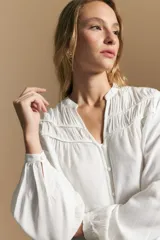 Camisa de manga larga color blanco, con cuello mao y escote en V. Presenta frunces en el canesú, cierre frontal con botones y mangas amplias con puños abotonados.