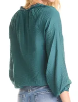 Blusa de satén color verde petróleo, con escote en V y cuello decorado con mostacillas bordadas. Presenta mangas largas con puños elásticos y ruedo también elastizado, brindando un ajuste cómodo y holgado.