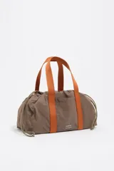 Mini bolso de mano Bimba y Lola confeccionado en nylon color marrón, con asas de cinta en color naranja y cierre mediante cordones ajustables en los laterales. Presenta logo metálico en la parte frontal.