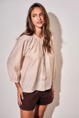 Blusa oversize de algodón y viscosa, color beige con rayas verticales finas. Presenta cuello redondo con frunces, cierre frontal con botones y mangas tres cuartos abullonadas con puños elásticos.