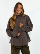 Camisa de algodón color gris oscuro, con cuello alto con volados, cierre frontal oculto y mangas largas abullonadas con puños con volados.