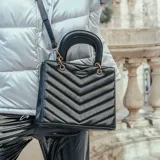 Cartera negra de símil cuero acolchado con diseño de líneas horizontales en forma de V. Tiene acceso con cierre y broches imantados, correa regulable, herrajes dorados y tres compartimentos internos. Se puede usar como bandolera o de mano.