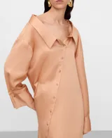 Vestido camisero midi de tejido satinado en tono salmón, con cuello italiano desestructurado, mangas largas con puños abotonados y una original botonadura frontal asimétrica.