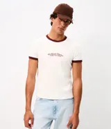 Remera estilo baby tee de color blanco crudo, con cuello redondo y ribetes en contraste de tono bordó. Presenta mangas cortas y una estampa frontal con la inscripción "Worldwide Rhythm".