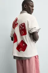 Camiseta blanca de corte relajado (relaxed fit) con cuello redondo y manga corta. Presenta un estampado grande en la espalda que simula dados rojos con letras y corazones.