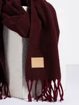 Bufanda de tejido suave en color rosado, con diseño liso y terminaciones con flecos en los extremos. Incluye una etiqueta de cuero sintético con el logo de la marca en uno de los bordes.
