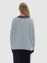 Buzo tejido de punto con diseño oversized, cuello en V profundo y mangas largas. Presenta un patrón de trenzas verticales en color verde oscuro con detalles en contraste de color rosado en el cuello, puños y ruedo.