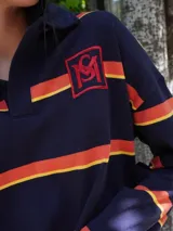 Buzo tipo rugby de algodón con diseño de franjas horizontales en tonos beige, negro y fucsia. Presenta cuello tipo polo en contraste y cierre frontal parcial.