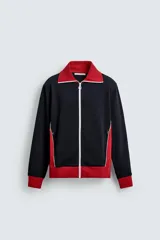 Chaqueta deportiva de corte regular, confeccionada en tejido piqué con interior forrado en red. Presenta un diseño color block en negro y rojo con detalles en blanco, cuello subido, cierre frontal con cremallera y bolsillos laterales con cierre.