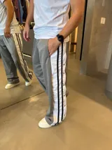 Pantalón deportivo de corte holgado en color gris, con franjas laterales blancas y negras que incluyen botones a presión. Presenta un diseño de estilo retro con caída amplia.