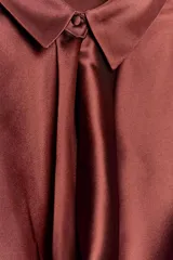 Blusa de tejido satinado color bordó, con cuello solapa, manga larga y detalle de abertura drapeada en el pecho. Cuenta con cierre frontal mediante un botón forrado.