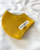 Tapabocas de tela color mostaza con elástico blanco y etiqueta con el logo de la marca Valente.