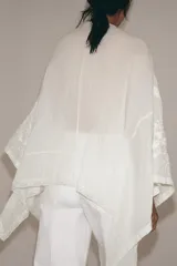 Blusa tipo capa de algodón con escote en pico y mangas largas amplias. Presenta bordados florales a tono en toda la prenda, bajo asimétrico y cierre frontal con botones.