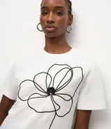 Remera de corte box, levemente amplia, confeccionada en algodón. Presenta cuello redondo, mangas cortas y un diseño de flor bordada en hilo negro en la parte delantera.