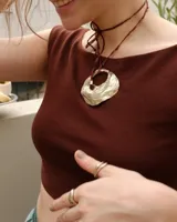 Collar artesanal con dije de bronce macizo en forma orgánica irregular, similar a una concha marina, suspendido de un cordón de seda natural negro. El diseño permite diversas formas de uso, ya sea ajustado al cuello o largo.