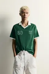 Polo de fútbol retro color verde oscuro, de corte regular fit, con cuello solapa blanco, mangas cortas con ribetes blancos y bandas en los hombros. Cuenta con estampado frontal del número 80 y un gráfico con efecto lavado.