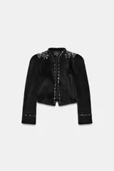 Chaqueta entallada negra de manga larga y cuello redondo, con apliques de tachas metálicas en los hombros, a lo largo de la abertura frontal y en los puños. Cuenta con cierre frontal con gancho metálico.