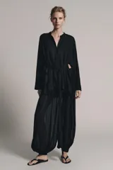 Camisa negra de cuello redondo y manga larga ancha con silueta fluida, con cierre frontal de botones y aberturas laterales en el bajo.