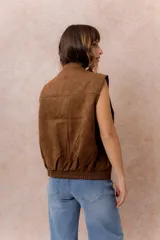 Chaleco de gamuza color marrón con acabado aterciopelado, cuello alto con broche a presión, cierre frontal metálico y bolsillos laterales con cierre.