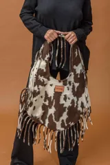 Bolso tipo hobo de gran capacidad con estampado animal print de vaca en tonos marrón y blanco. Presenta una etiqueta de cuero con el logo de la marca en el frente y un diseño de caída relajada.
