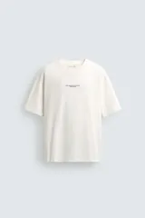 Camiseta de corte relajado, color crudo, con cuello redondo y manga corta. Presenta un diseño con texto estampado y bordados a contraste en la parte frontal.