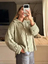 Saco tejido en color verde oliva con diseño de ochos y trenzas. Presenta cuello estilo camisero, cierre frontal con botones tipo joya, mangas holgadas y silueta oversize.