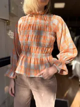 Blusa de viyela con estampado a cuadros en tonos naranja y celeste. Presenta un diseño con frunces en el cuerpo, cuello alto, mangas largas abullonadas con puños elásticos y terminación con volados.