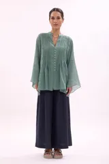 Camisa de corte holgado y diseño fluido, confeccionada en tela semitransparente de color verde. Presenta cuello mao, cierre frontal con botones pequeños y detalles de alforzas verticales en el frente. Las mangas son largas y acampanadas, aportando un estilo ligero y sofisticado.