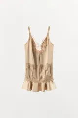 Vestido mini de tejido satinado color beige, con escote en pico adornado con encaje, tirantes anchos y detalle de encaje bordado en la cintura. El bajo presenta un acabado con volantes.