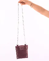 Porta celular de cuero natural color bordó, con diseño rectangular y solapa de cierre. Presenta detalles de tachas metálicas plateadas en los laterales y correa de cadena metálica.