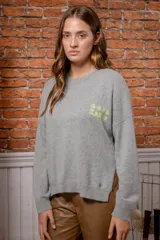 Sweater de punto color gris con bordado en color verde neón en el pecho con la frase 'GOOD KARMA'. Presenta cuello redondo, mangas largas y un diseño con tajos laterales en el ruedo.