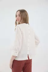 Blusa de lino color marrón, de corte recto y mangas 3/4. Presenta escote redondo con pequeños volados y pliegues frontales, además de detalles de encaje en las mangas y puños con volados.
