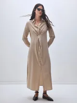 Vestido camisero largo color beige, con cuello clásico, mangas largas y cierre frontal con botones. Presenta un nudo decorativo a la altura de la cintura y una abertura en el ruedo.