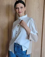 Camisa blanca de manga larga con cuello alto fruncido y botones marrones en el frente. Presenta paneles laterales y mangas en color beige.