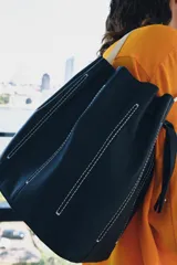 Bolso de hombro tipo saco confeccionado en piel azul marino, con detalle de pespuntes blancos verticales, asas finas de hombro en color blanco y cierre fruncido.