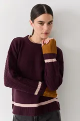 Buzo de punto color crudo con mangas de textura trenzada, detalles en contraste en los puños y franjas horizontales en el ruedo. Presenta cuello redondo y un pequeño bordado en el pecho.