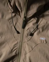 Campera rompevientos Obey de color tostado, con capucha ajustable, cierre frontal completo y bolsillos laterales con cierre. Presenta logo estampado en el pecho y cordones ajustables en el ruedo.