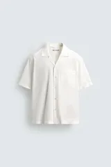 Camisa de calce relajado confeccionada en tejido ligero de algodón con textura. Presenta cuello bowling, manga corta, un bolsillo de parche en el pecho y cierre frontal con botones. El bajo cuenta con aberturas laterales.