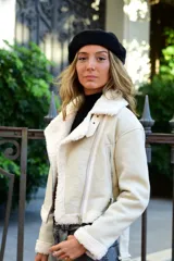 Campera de efecto cuero color blanco off-white con interior y terminaciones de corderito sintético. Presenta cierre frontal cruzado, cuello amplio con solapas, bolsillos con cremallera y cinto ajustable en la parte inferior.