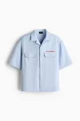 Camisa de manga corta color celeste, de corte relajado, con cuello abierto, bolsillos delanteros con solapa y cierre de botones. Cuenta con detalle de texto bordado en rojo sobre el bolsillo superior.