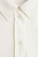 Camisa de manga larga color blanco hueso, con cuello camisero clásico y cierre frontal de botones. Presenta un corte recto y un bajo redondeado.