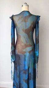 Vestido largo de microtul translúcido con estampado floral en tonos azules y marrones. Posee escote en V con lazo ajustable, mangas largas acampanadas con volados y una abertura lateral en la falda.